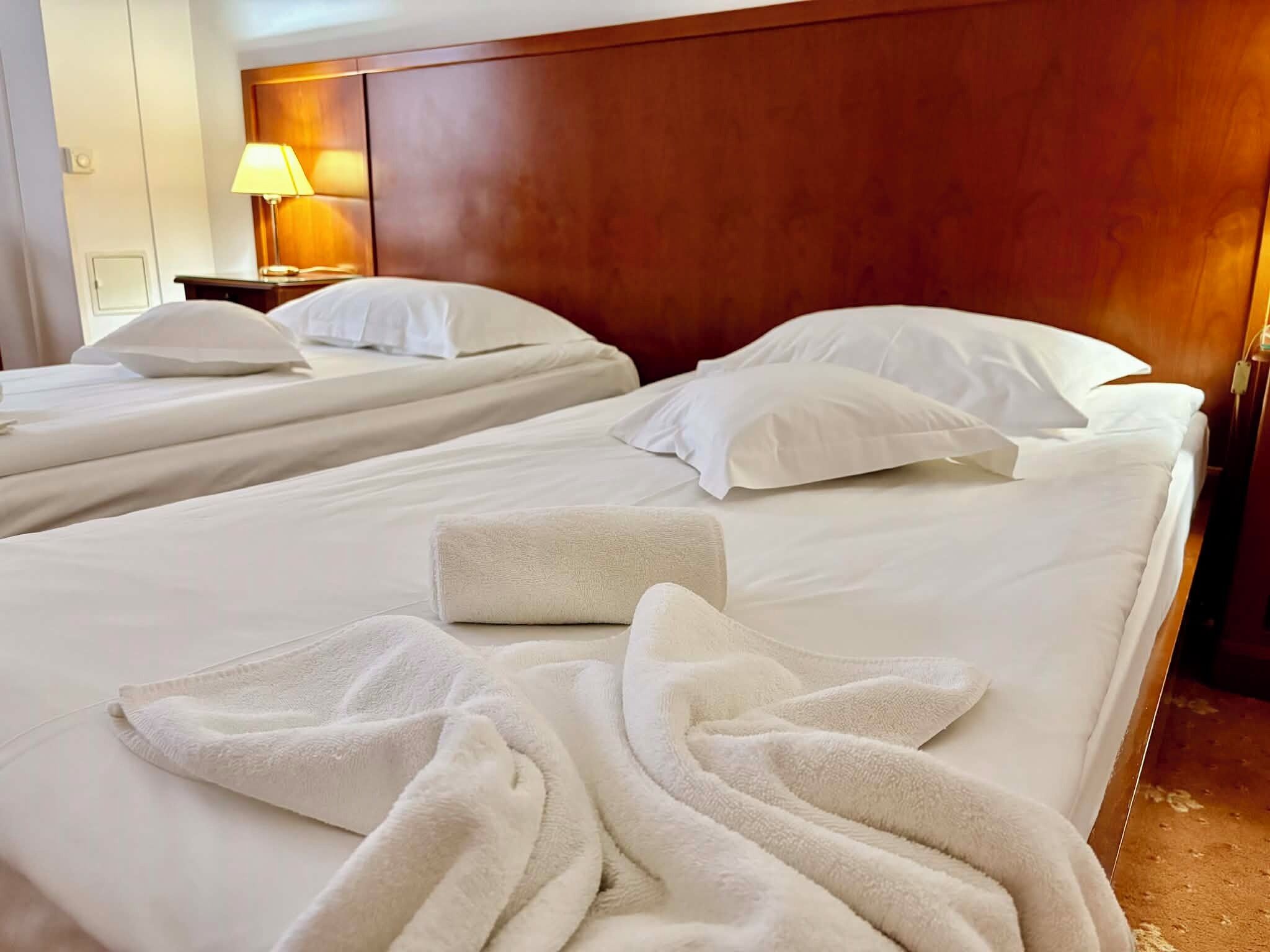 Ein Hotelzimmer mit zwei Betten, jeweils mit weißen Bettwäsche und Kissen, einem hölzernen Kopfteil, einem Nachttisch mit Lampe und einem Handtuch auf dem Bett.