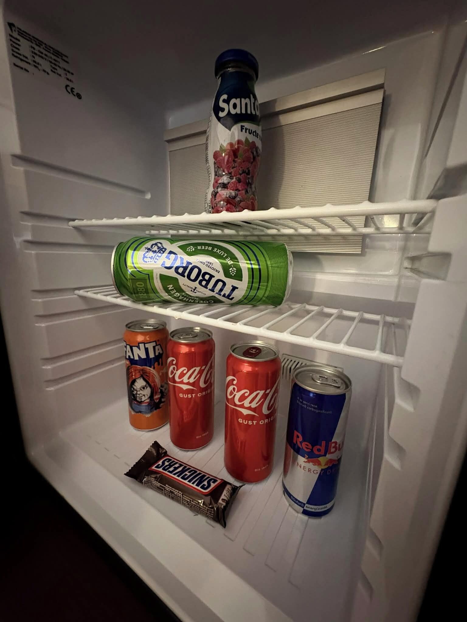 A refrigerator with a bottle of Santa Cruz Fructis, a can of 1880, and several cans of Coca-Cola and Red Bull.