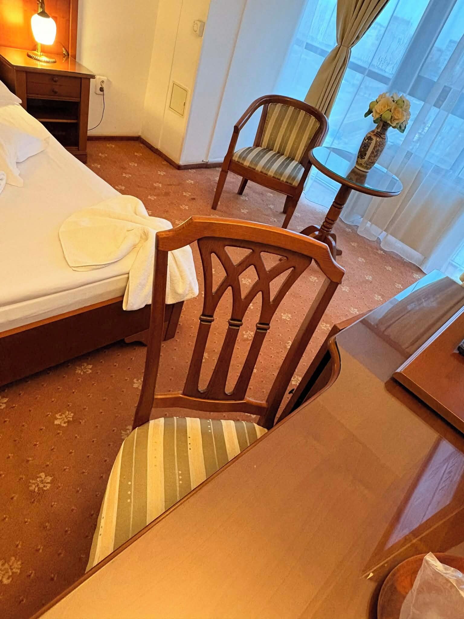 Una habitación de hotel con una cama, una silla de madera, una mesita redonda con un florero y una lámpara en una mesita de noche.