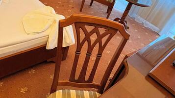 Una habitación de hotel con una cama, una silla de madera, una mesita redonda con un florero y una lámpara en una mesita de noche.