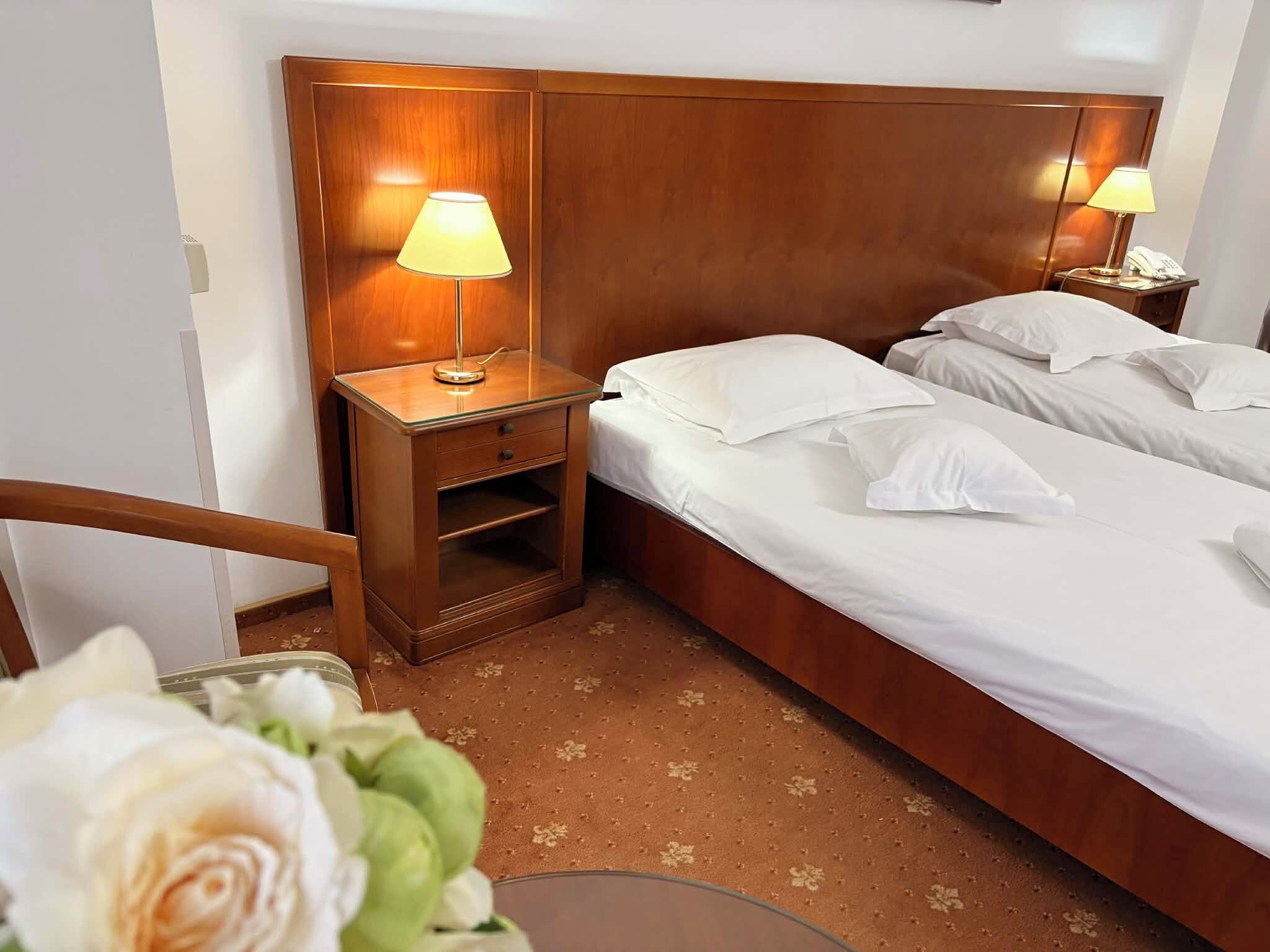 Habitación de hotel con dos camas individuales, cabecera de madera, lámparas de noche, un cajón pequeño y un arreglo floral.