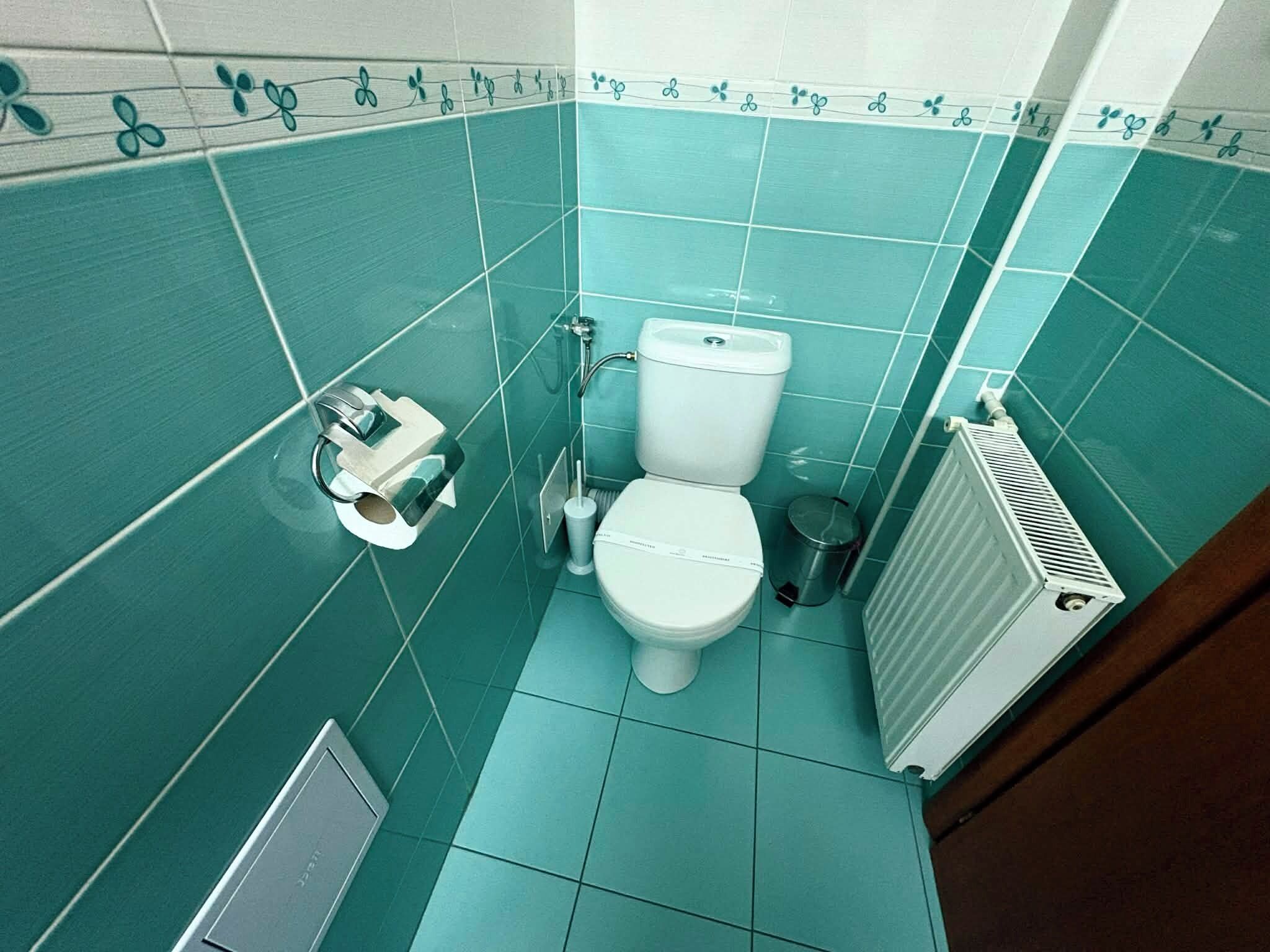 A bathroom with teal tiles, a toilet, a bidet, a trash can, and a radiator.