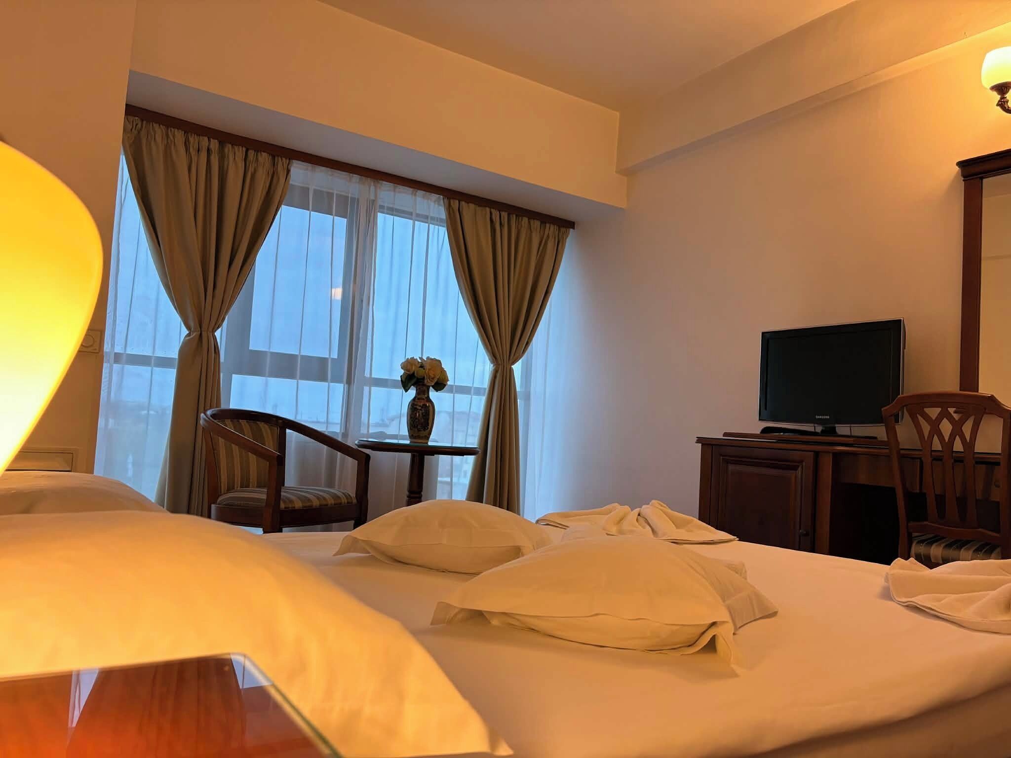 Ein Hotelzimmer mit einem Bett, einem Schreibtisch mit Fernseher, einem Sessel und einem Fenster mit Vorhängen.