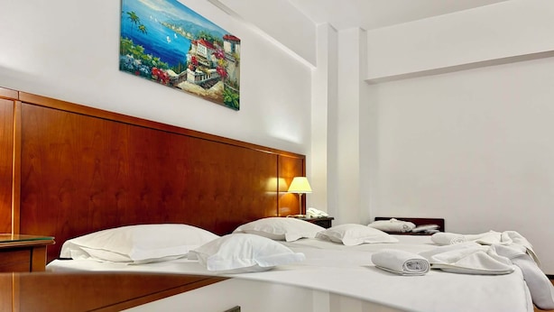 A hotel room with a wooden headboard, a painting of a coastal scene, a bed with white linens, and a bedside table with a lamp and phone.