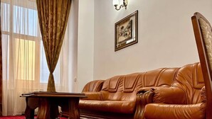 A room with a brown leather sofa, a wooden coffee table, a red carpet with a floral pattern, a window with sheer curtains, and a wall-mounted picture frame.