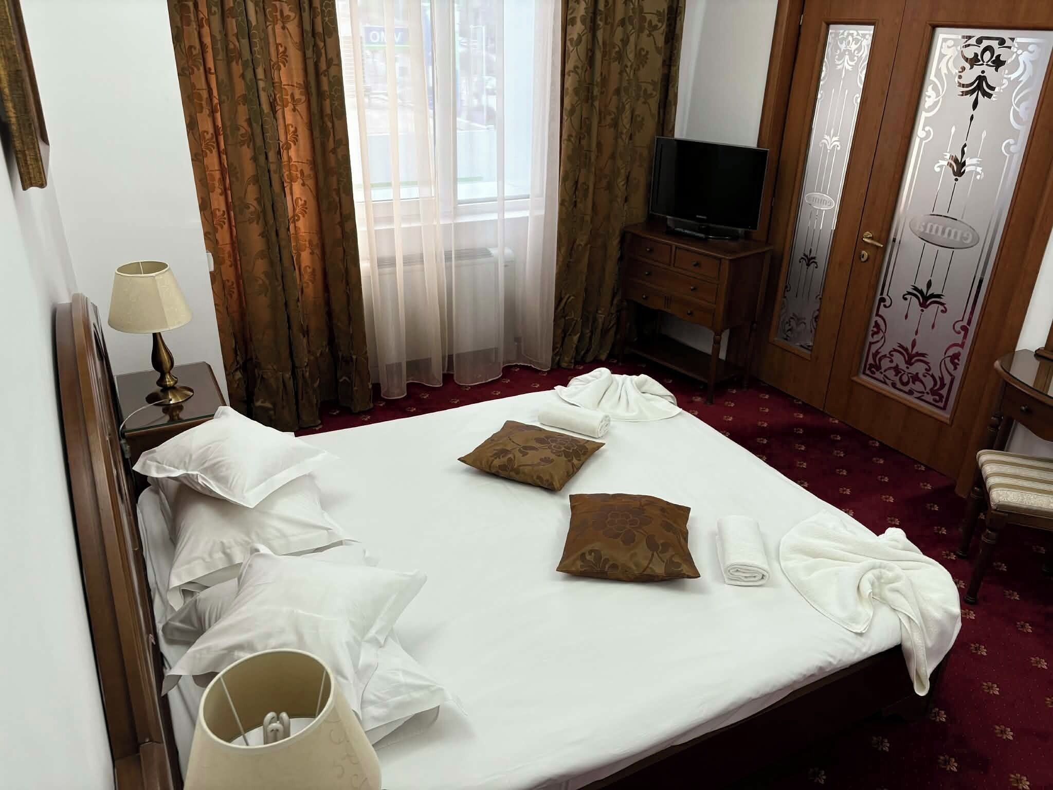 Een hotelkamer met een bed, kussens, een televisie, een lamp, een kledingkast en een raam met gordijnen.