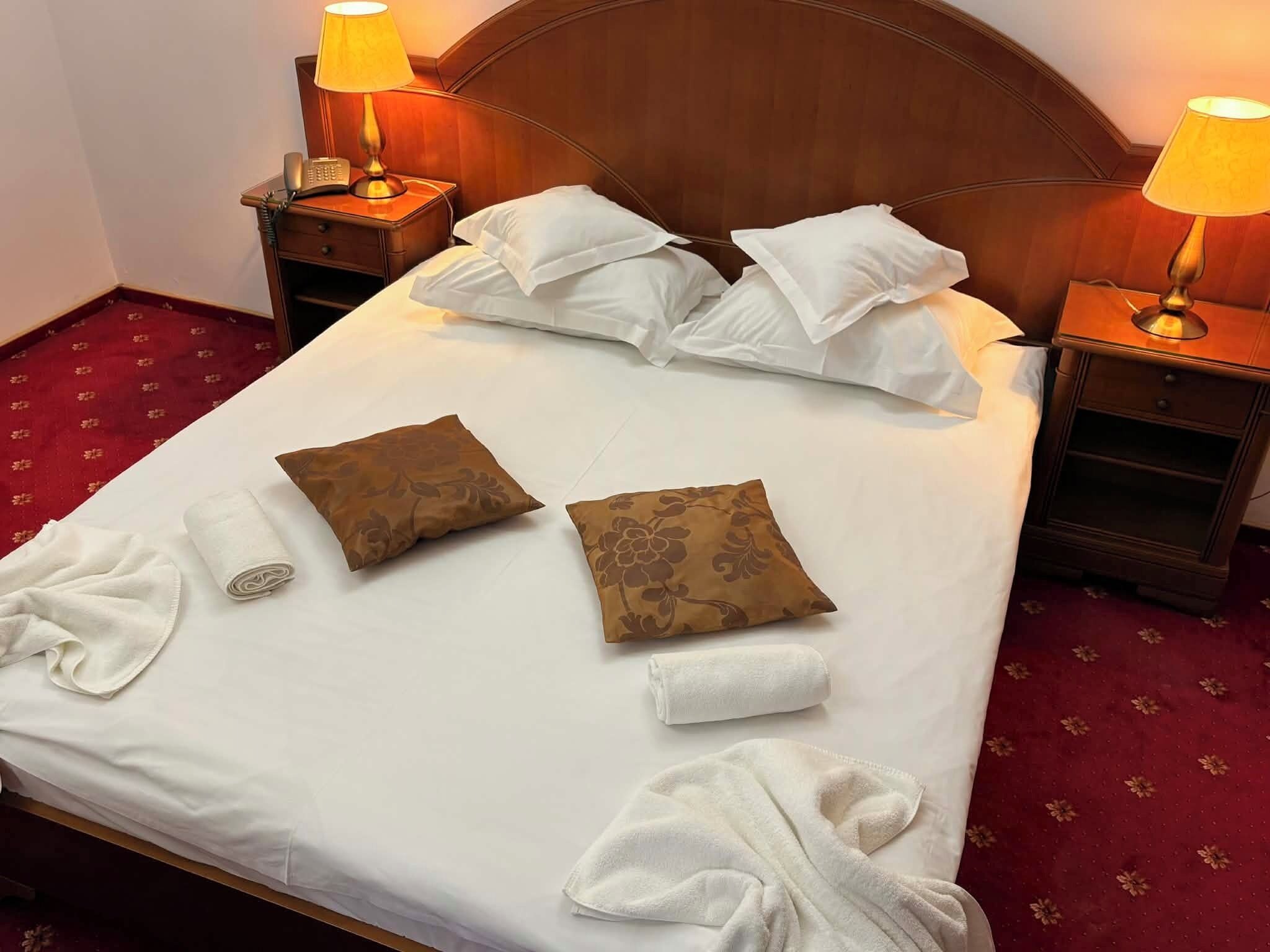 Ein ordentlich bezogenes Bett mit weißen Leinentüchern, zwei braunen Kissen mit Muster und zwei aufgerollte weiße Handtücher. Auf den hölzernen Nachttischen stehen jeweils zwei Nachttischlampen.