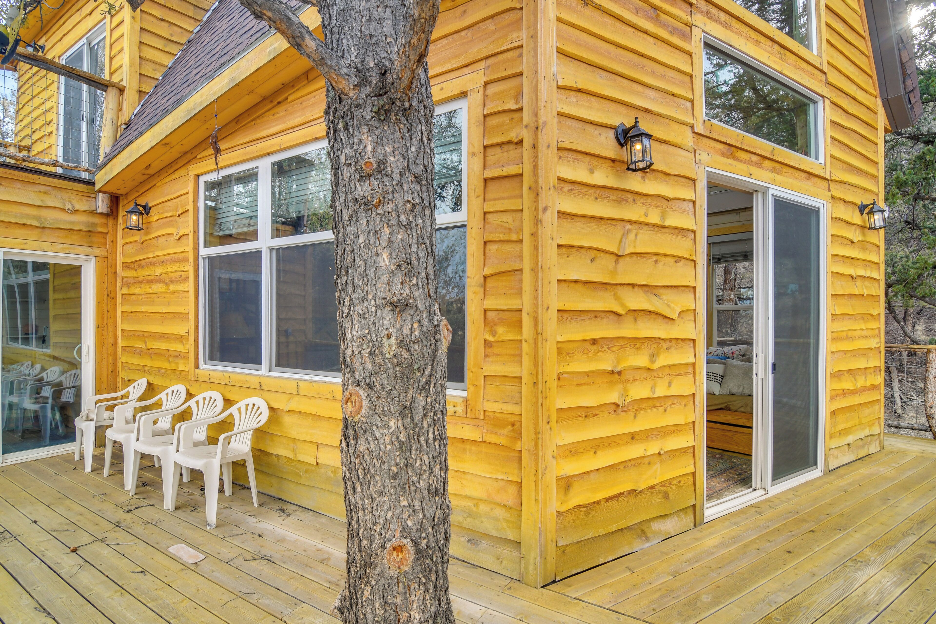 Une cabane en rondins jaune avec une terrasse, des chaises blanches et un grand arbre devant.