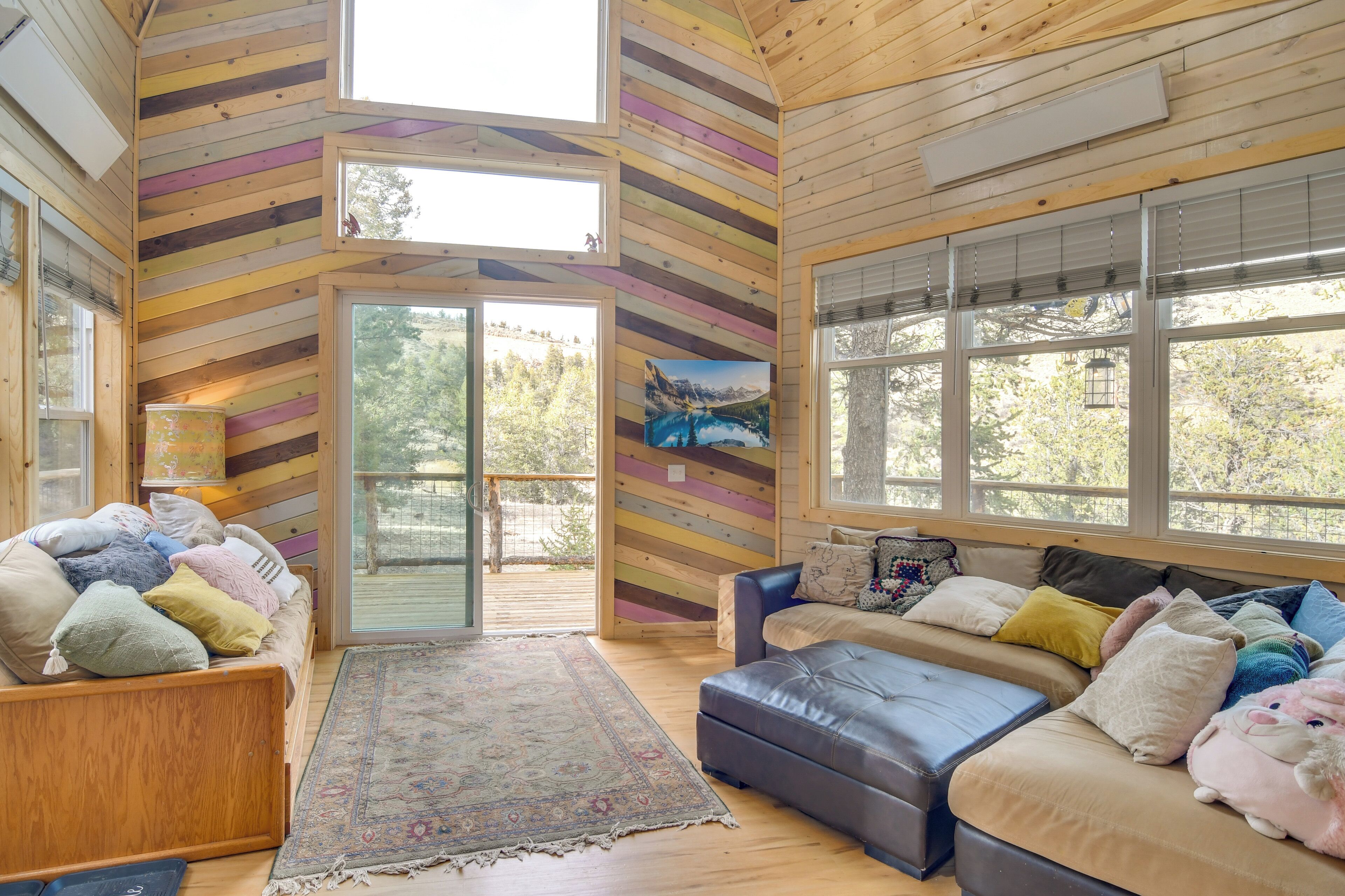 A cozy cabin interior with a wooden slat wall, a large window, a sofa, and a bed.