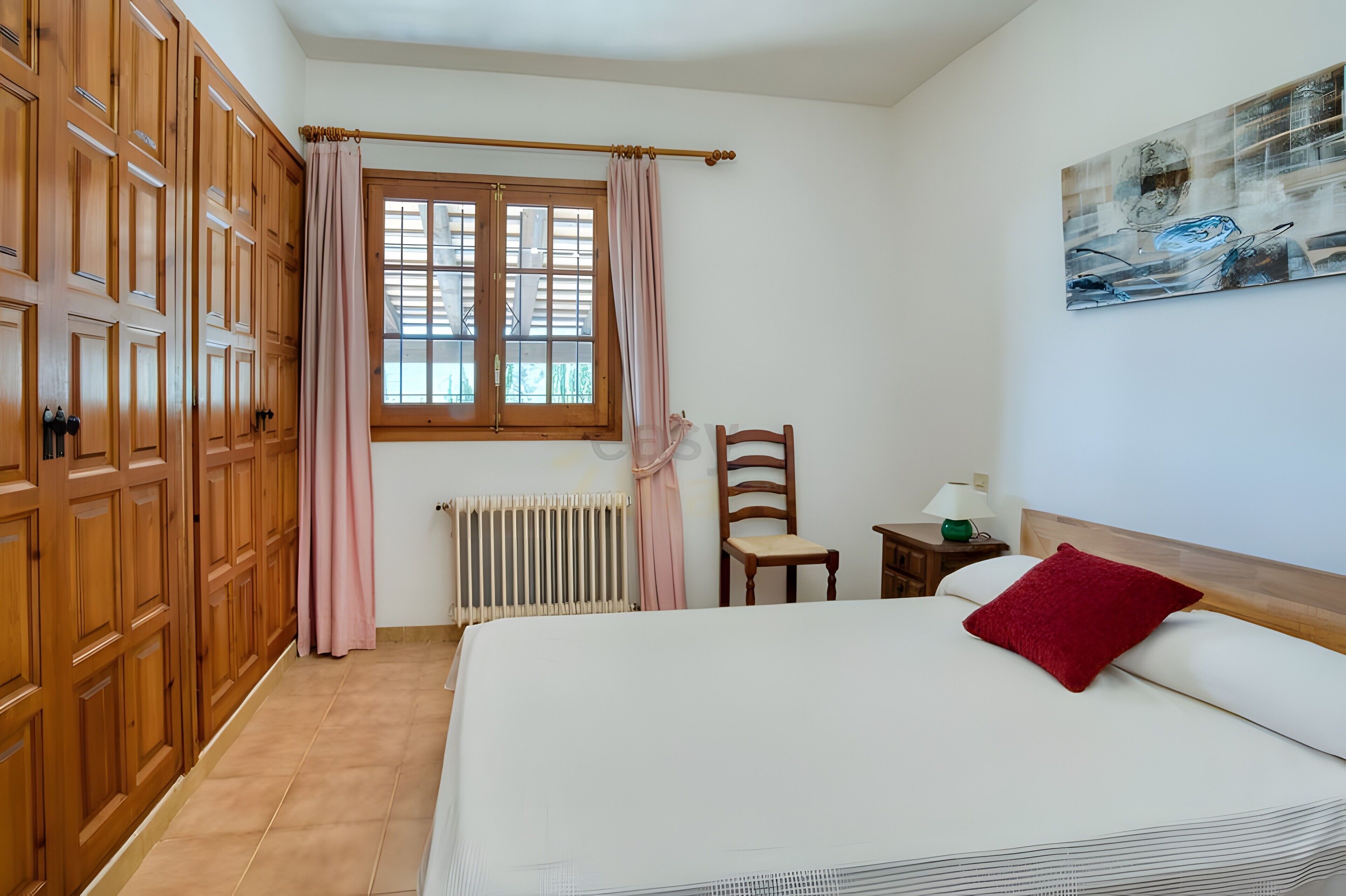 Une chambre à coucher avec un lit, une chaise, un radiateur, une fenêtre avec des rideaux et un tableau au mur.