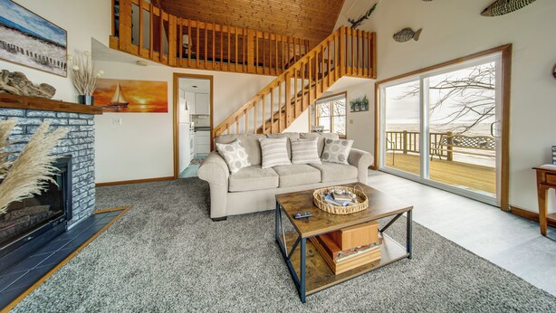 A spacious living area with a fireplace, a wooden staircase, a large window with a view, and a cozy seating area. - Lakeside Living on Wine Trail! Geneva Home w/ Deck (Geneva)