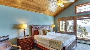 A bedroom with a wooden ceiling, a bed with a headboard, two bedside tables with lamps, a chair, and a window with a view.