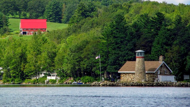 Un faro de piedra junto a un lago, rodeado de un bosque frondoso y un granero rojo al fondo.