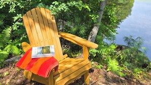 Una silla Adirondack de madera con un libro titulado 'This is Happiness' de Niall Williams, ubicada en un entorno boscoso junto al lago.