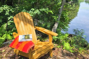 A wooden Adirondack chair with a book titled 'This is Happiness' by Niall Williams, placed on a forested lakeside setting. - Waterfront Home w/ Access to Lake Anasagunticook! (Canton)