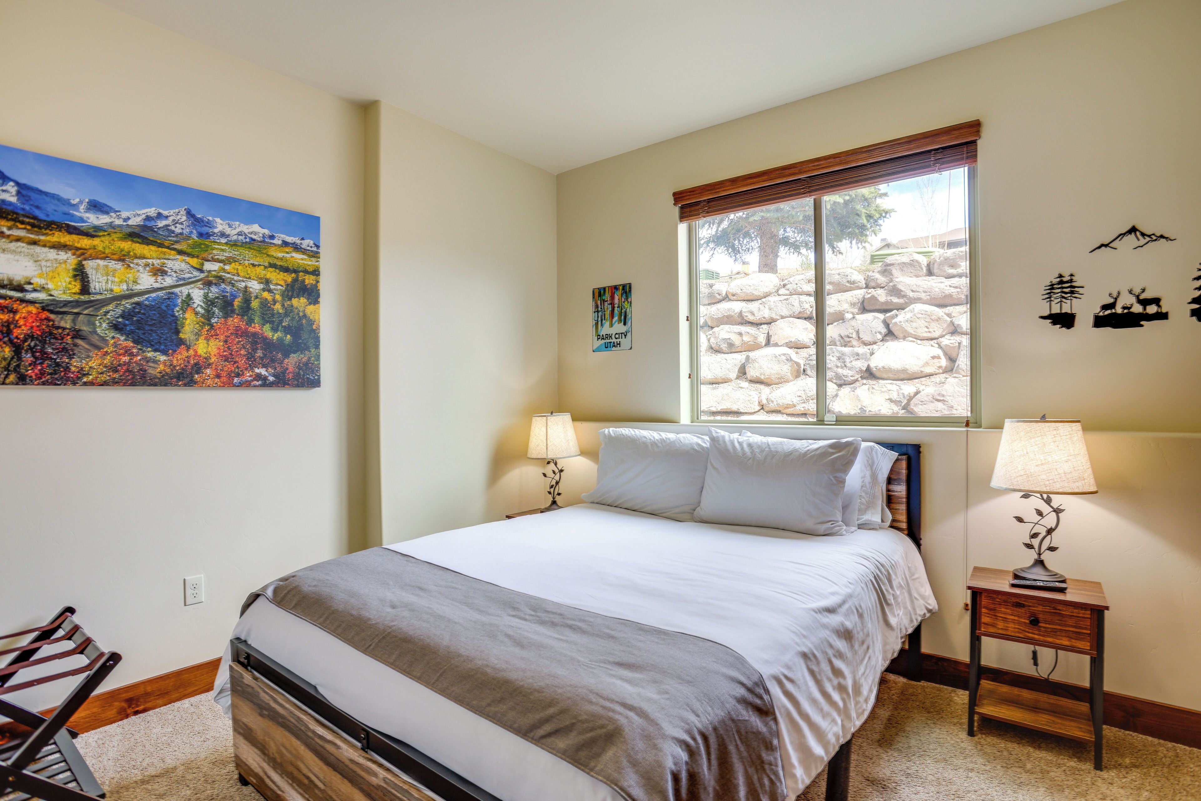 A bedroom with a bed, two bedside lamps, a window with a stone wall, a painting, and a wall-mounted decoration.