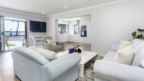 Una sala de estar moderna con un sofá, sillones y una mesa de centro. Hay una zona de cocina con armarios y un televisor montado en la pared.