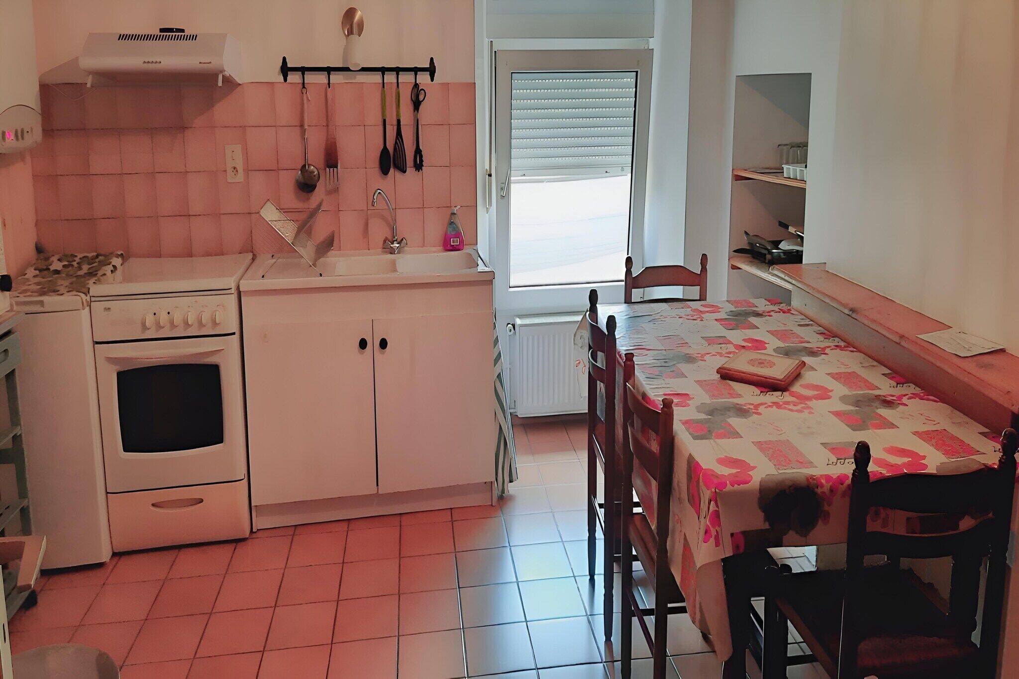 A kitchen with a stove, oven, sink, and hanging utensils. There is a dining table with chairs and a window with blinds.