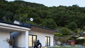 Ein modernes Haus mit einem gepflasterten Weg, der zum Eingang führt, umgeben von üppiger Vegetation und einer hügeligen Landschaft im Hintergrund.