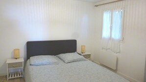 Ett sovrum med en säng, två sängbord, ett fönster med gardiner, ett element och en taklampa.