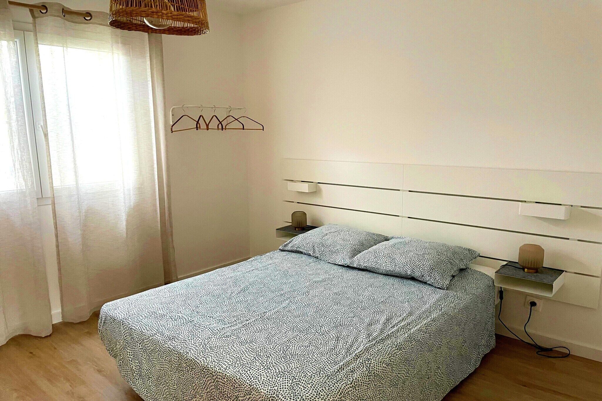 A bedroom with a bed, a bedside table, a wall-mounted shelf, and a window with curtains.
