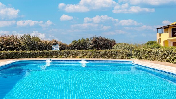 A clear blue swimming pool with a tiled edge, surrounded by a hedge and a building with a yellow facade. - Holiday home with air conditioning and shared pool (Viddalba)