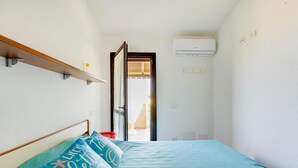 A narrow room with a bed, a shelf, an air conditioner, and a door leading to another room. - Holiday home with air conditioning and shared pool (Viddalba)