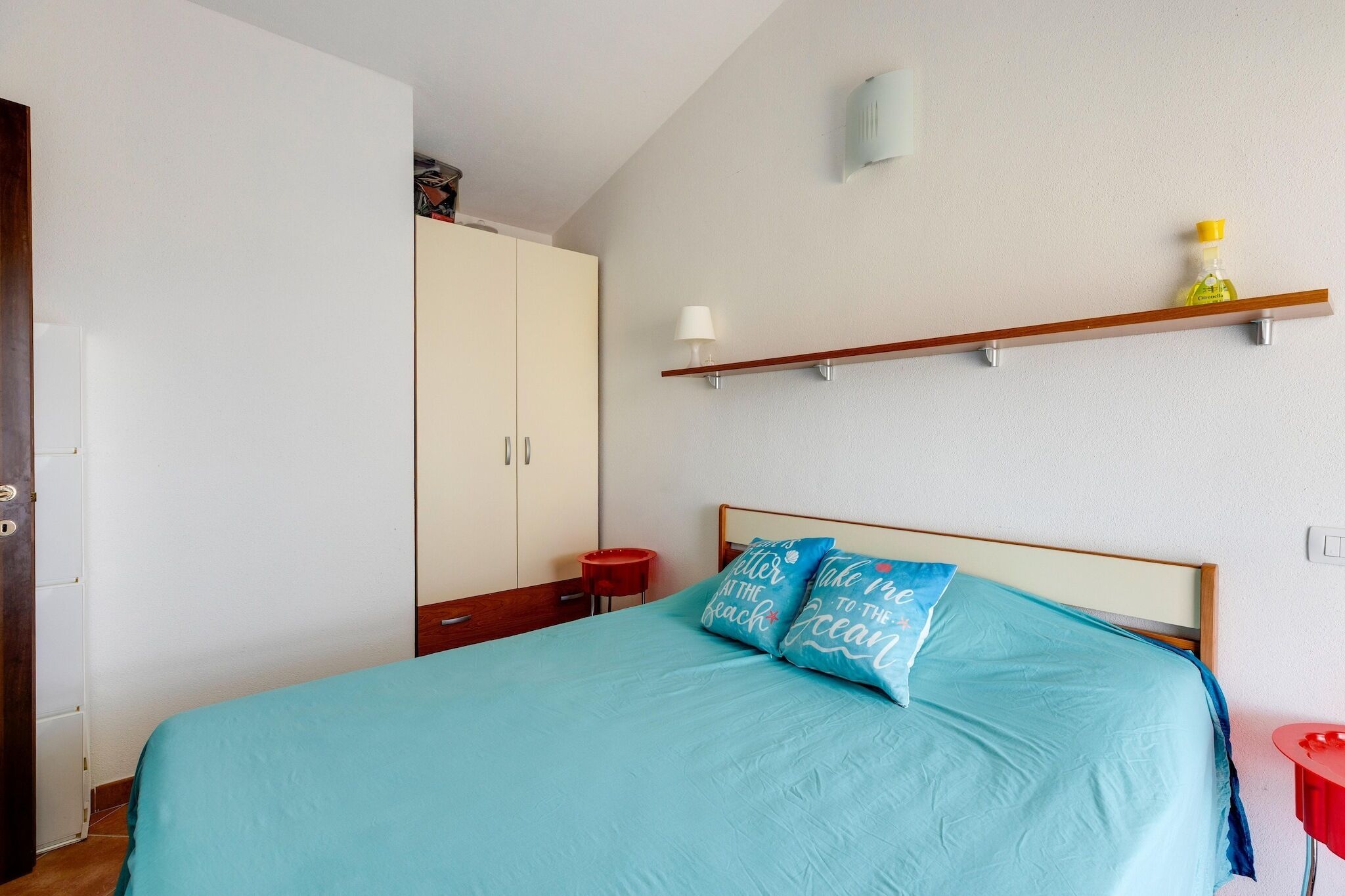 Un dormitorio con cama, armario, estante con lámpara y una mesita.