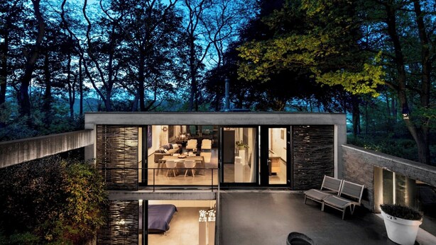 Uma casa moderna com fachada de vidro, um pátio com assentos e vista para a floresta ao redor.