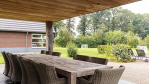 An outdoor patio with a wooden table and wicker chairs, a covered area, and a view of a brick building and greenery. - Achterhoek Villa with Heated Pool (Aalten)
