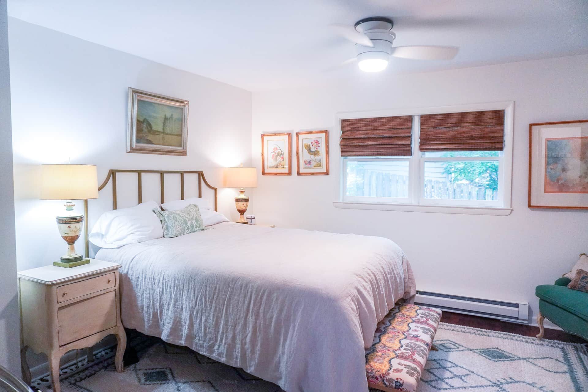 A bedroom with a bed, bedside lamps, a window with blinds, a ceiling fan, and a framed picture on the wall.