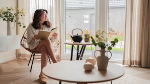 A woman sitting on a wicker chair reading a book, with a table and potted plants in a bright room with a view of a house through a glass door. - Modern Dune Retreat with Garden (Sint Maarten)