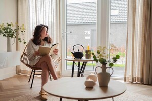 A woman sitting on a wicker chair reading a book, with a table and potted plants in a bright room with a view of a house through a glass door. - Modern Dune Retreat with Garden (Sint Maarten)