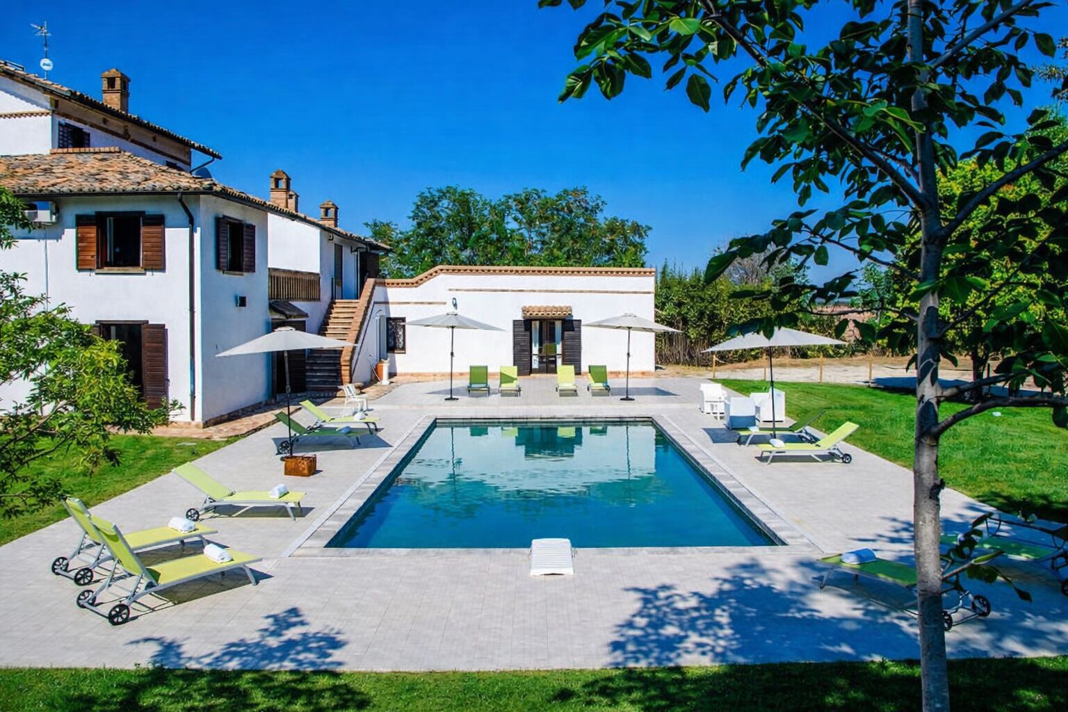 A swimming pool with lounge chairs and umbrellas, a white building with a tiled roof, and a clear blue sky.