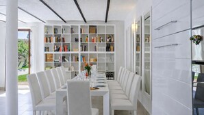 A dining area with a long table, white chairs, and a bookshelf filled with books and bottles.