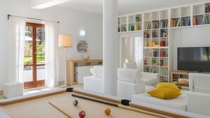 A modern living room with a pool table, white sofas, a bookshelf, and a television.