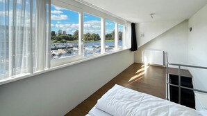 Een kamer met een groot raam dat uitkijkt op een jachthaven, een bed met wit beddengoed, een radiator en een metalen trap.