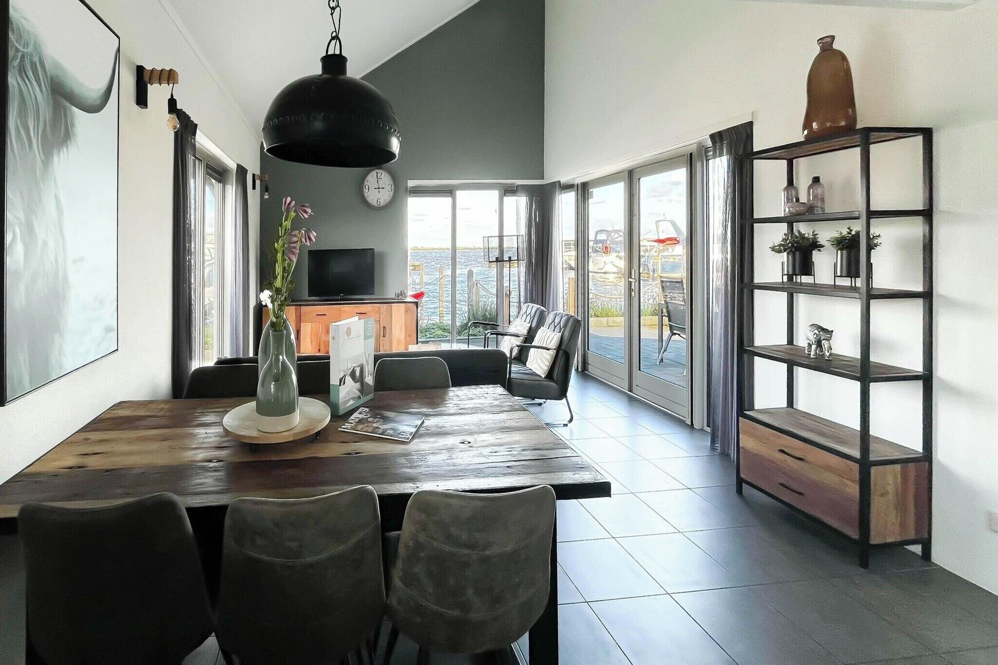 A spacious dining area with a wooden table and chairs, a large hanging light fixture, a television, and a view of the outdoors through large windows.