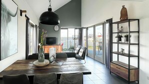 Een ruime eetruimte met een houten tafel en stoelen, een grote hanglamp, een televisietoestel en uitzicht op de omgeving door grote ramen.
