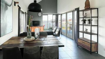 A spacious dining area with a wooden table and chairs, a large hanging light fixture, a television, and a view of the outdoors through large windows.