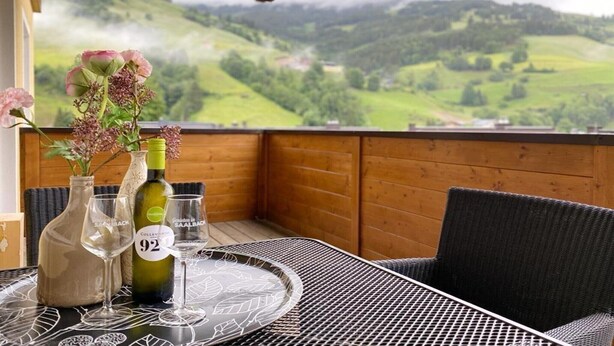 Ein Tisch mit einer Weinflasche, zwei Gläsern und einer Blumenvase auf einem Balkon mit Blick auf eine grüne Landschaft.