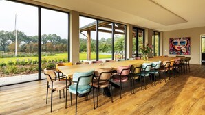 A modern dining area with a long wooden table and colorful chairs, large windows offering a view of a garden. - Villa with Heated Pool, Sauna (Aalten)