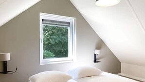 A bedroom with a sloped ceiling, a window with a view of trees, two bedside lamps, and a bed with white bedding. - Modern Coastal Retreat with Garden (Sint Maarten)