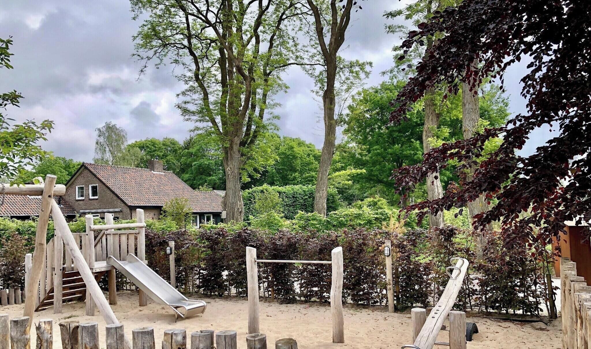 Un terrain de jeux avec un toboggan et des structures d’escalade, entouré d’arbres et avec une maison en arrière-plan.