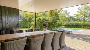 Un patio al aire libre con una mesa de madera y sillas de mimbre, una piscina y vistas a un jardín.