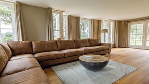 Un salón amplio con un sofá seccional de cuero marrón, una mesa redonda y ventanas grandes con cortinas.