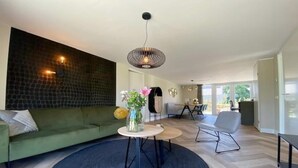 Una sala moderna con un sofá verde, una mesa redonda con un florero, una lámpara colgante y una zona de comedor al fondo.