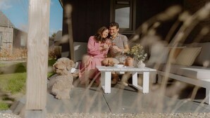 Una pareja sentada en un sofá blanco al aire libre, con una pequeña mesa entre ellos, un perro y una estructura de pérgola de madera.