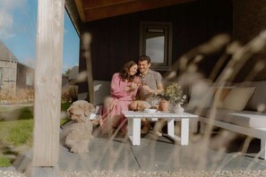 A couple sitting on a white outdoor sofa with a small table between them, a dog, and a wooden pergola structure. - Coastal Home with Garden for 6 (Sint Maarten)