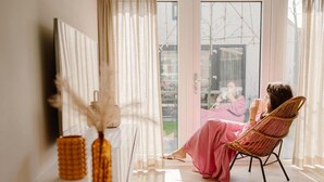 Una habitación con un ventanal grande, una silla de mimbre y un florero con plantas secas.